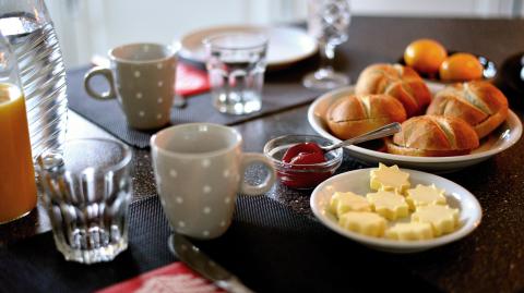 Das Foto zeigt eine gedeckten Tisch mit Frühstück. Es besteht aus je einer Karaffe mit Wasser und Orangensaft, mehreren Gedecken aus Becher, Gläser, Teller, Besteck auf Tischsets und Butter, Brötchen, Eiern und Marmelade.