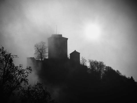 Ein Foto in schwarz-weiß der Burg Trifels, die im Nebel liegt und die Sonne leicht durchscheint.